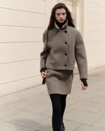 Model walking outdoors wearing a textured taupe wool jacket and skirt set with a dark shoulder bag