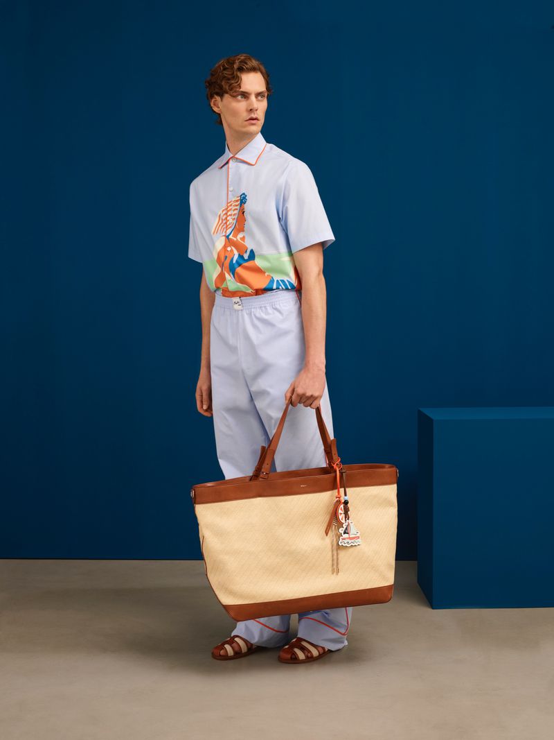 Male model wears a patterned shirt, blue pants, and sandals while holding a large tote bag against a blue backdrop.