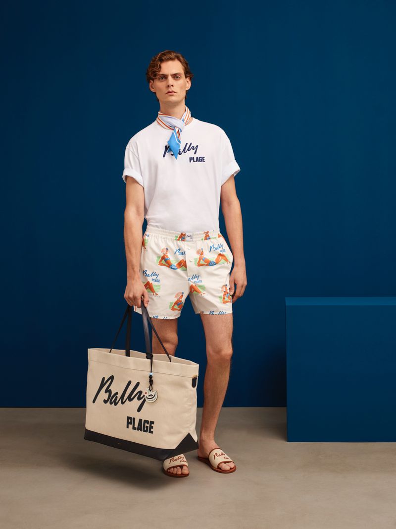 A stylish male fashion model wears printed shorts and a white tee, holding a Bally tote bag in a studio shot against a blue background.