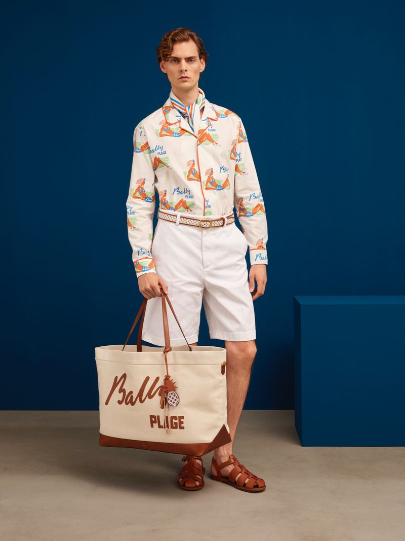 A male fashion model in a patterned button-down shirt, white shorts, and sandals holding a tote bag against a dark blue background.