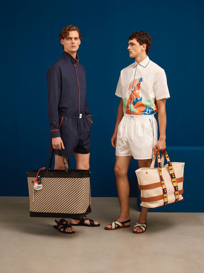 Two adult men pose indoors in fashion apparel, including short-sleeve and long-sleeve shirts, shorts, sandals, and tote bags on a blue background.