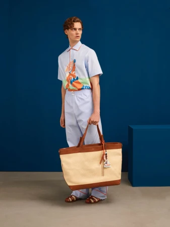 Male model wearing light blue patterned shirt and trousers holding a large canvas tote bag in a studio.