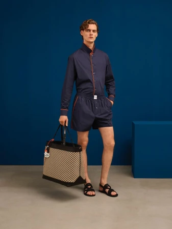Male model in navy shirt and shorts holding a striped tote bag against a blue and grey studio background for Bally S/S 26