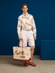 Male model wearing patterned shirt and white shorts holding a large canvas tote bag in a studio with a blue backdrop.