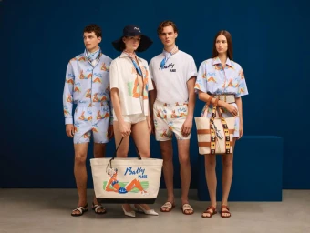 Four fashion models posing in matching resort wear and carrying large canvas tote bags against a solid dark blue studio backdrop.