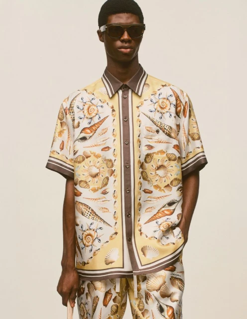 Fashionable adult man wearing a sea shell-patterned shirt and matching shorts with sunglasses indoors, posing for apparel campaign.