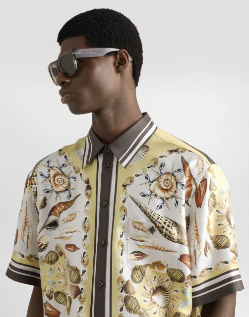 Dark-skinned man in a patterned short-sleeved shirt and sunglasses stands indoors against a neutral background in a portrait shot.
