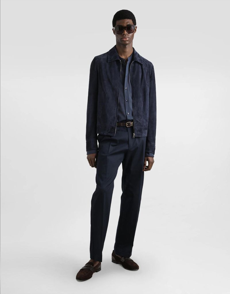 A black male model posing in a blue jacket, dress shirt, and pants, with brown loafers and sunglasses, against an isolated white backdrop.