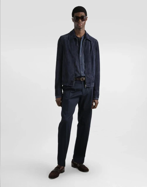 A black male model posing in a blue jacket, dress shirt, and pants, with brown loafers and sunglasses, against an isolated white backdrop.