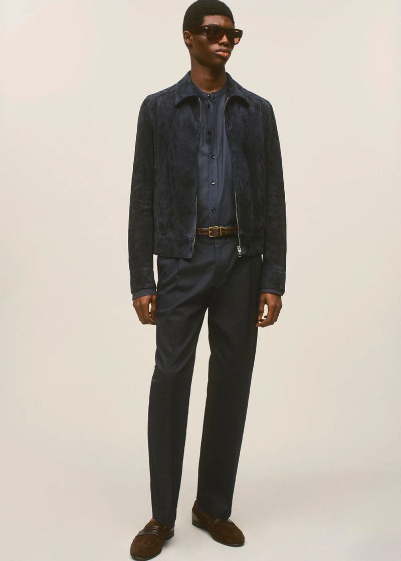 A full-length studio shot of a Black male model wearing a dark blue suede jacket, button-down shirt, dark trousers, brown belt, and loafers.