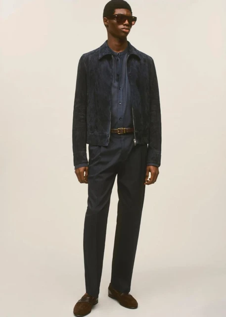 A full-length studio shot of a Black male model wearing a dark blue suede jacket, button-down shirt, dark trousers, brown belt, and loafers.