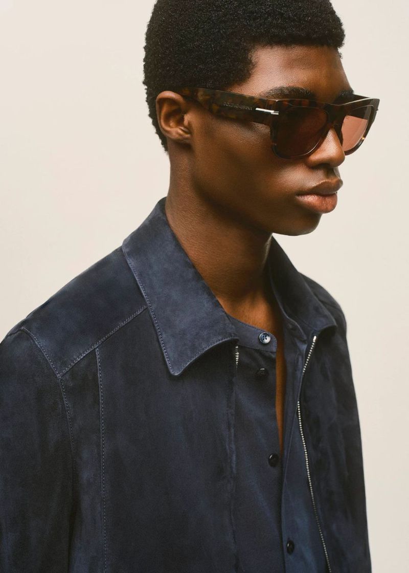Close-up of an adult man wearing sunglasses and a blue suede jacket, posing in front of an off-white background in a studio setting.
