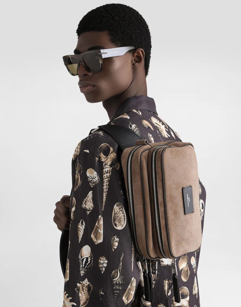Black man in graphic button-down shirt wearing sunglasses and carrying a brown suede backpack, posing against a neutral background.
