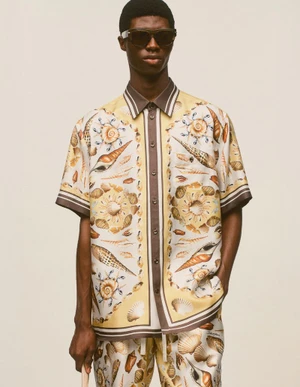 Model wearing a printed shell-pattern short-sleeve shirt and dark sunglasses against a neutral background