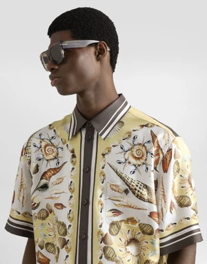 Model wearing a patterned short-sleeve shirt and dark sunglasses against a seamless white background.