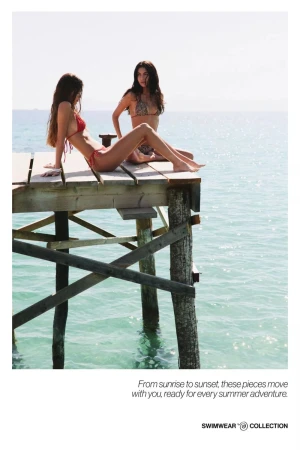 Two women in swimsuits pose on a wooden dock overlooking the ocean on a bright day in a swimwear fashion shot with a positive mood.
