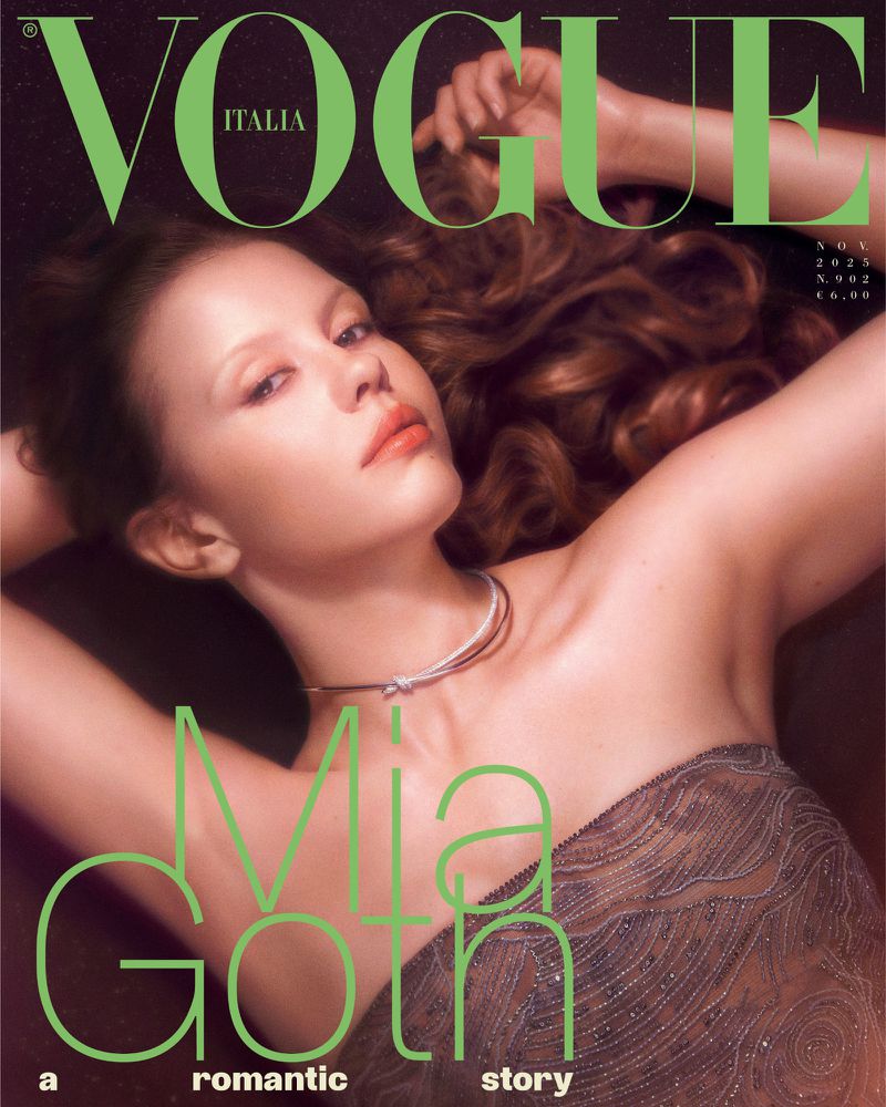 A woman with long wavy hair and a sheer top lies posing on a dark background for a magazine cover.