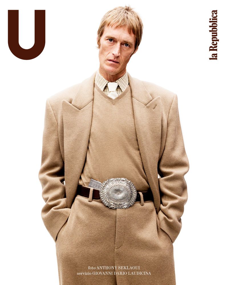 Editorial fashion shot of a blonde adult male model wearing a tan suit and silver belt buckle against a white backdrop for la Repubblica.