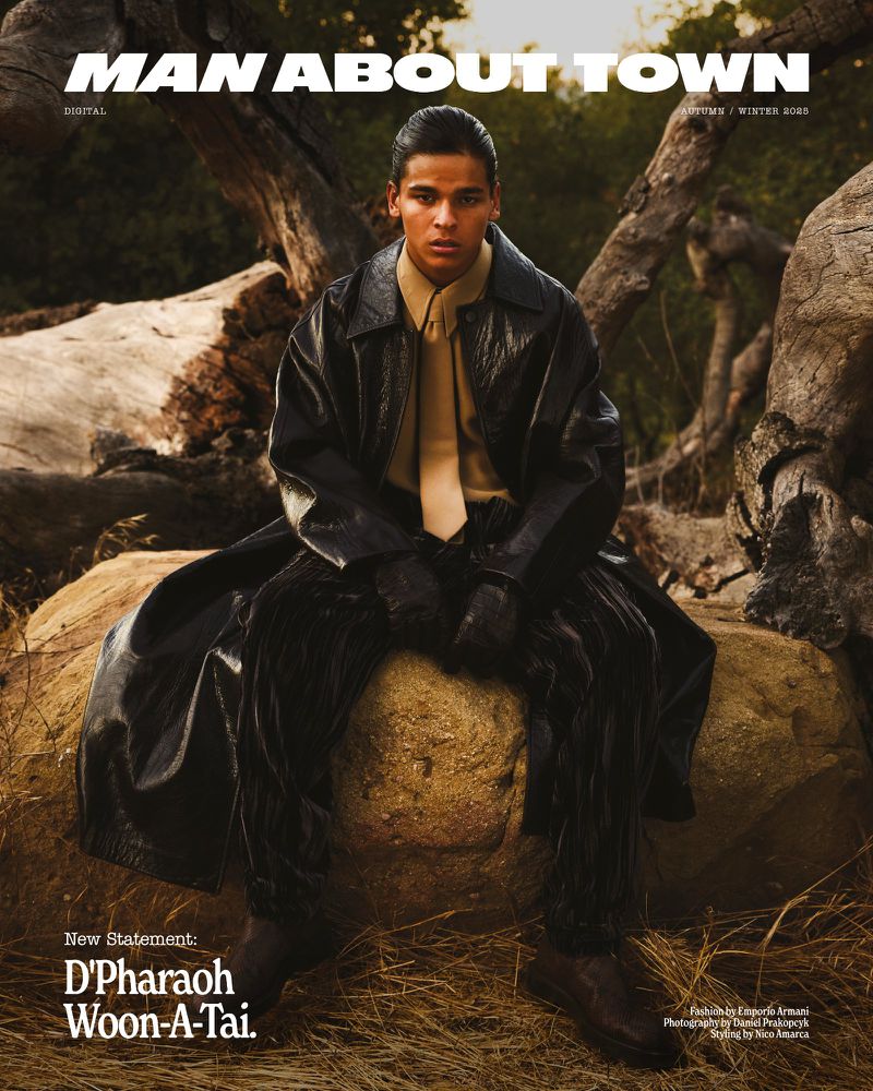 A male fashion model poses outdoors, seated on a rock, wearing a black vinyl trench coat, tan collared shirt and tie, and dark trousers.