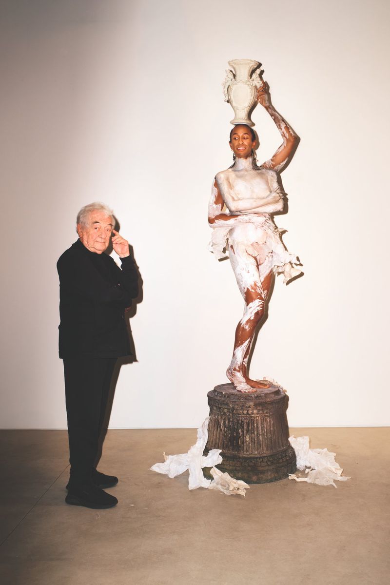 Elderly man in black suit standing next to a light-skinned model covered in white paint holding a vase on her head in a studio.