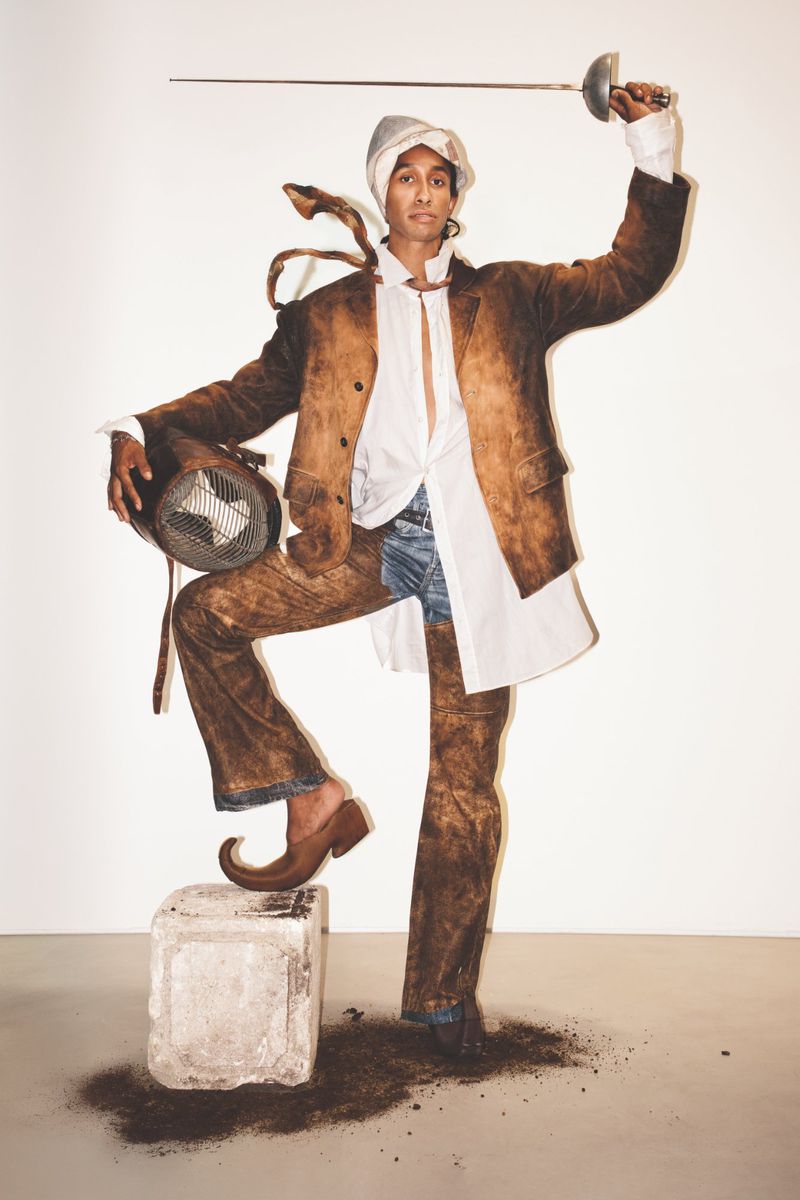 Fashion-forward man in a brown suit and turban poses with a fencing foil and mask in a photo studio, blending formal and avant-garde styles.