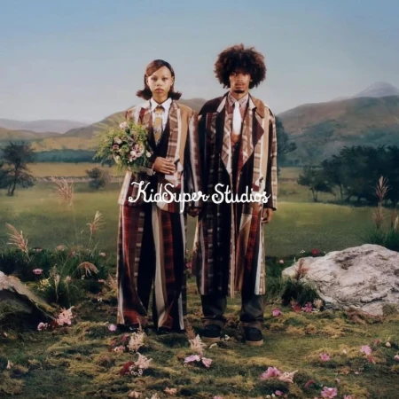 A stylish couple in matching striped outfits stands in a field with rolling hills, wearing suits and holding flowers, in a natural setting.