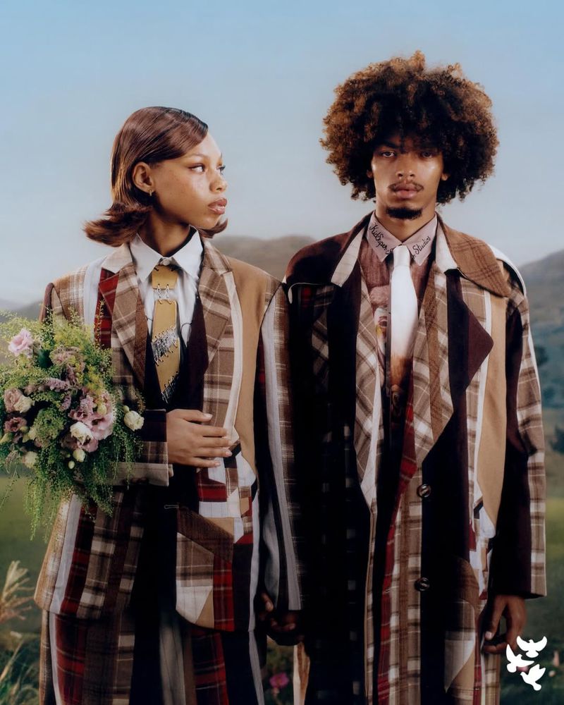 Matching couple wearing patchwork plaid suits in an outdoor setting, holding flowers, with a mountainous backdrop and soft natural light.