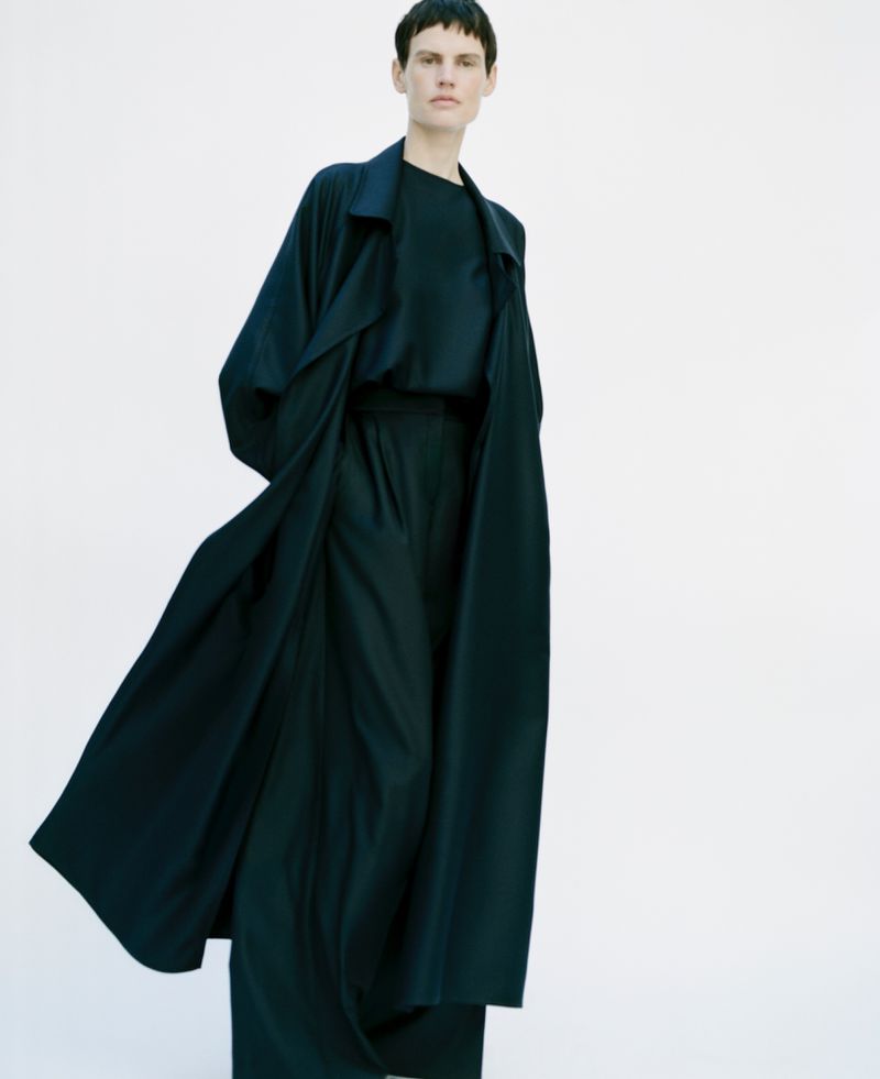 A woman wearing a dark, flowing trench coat and matching pants stands against a bright white background in a fashion editorial shot.