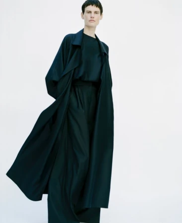A woman wearing a dark, flowing trench coat and matching pants stands against a bright white background in a fashion editorial shot.