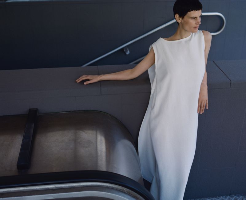 Model Saskia de Brauw in a white sleeveless dress leaning against a dark architectural wall for Dušan