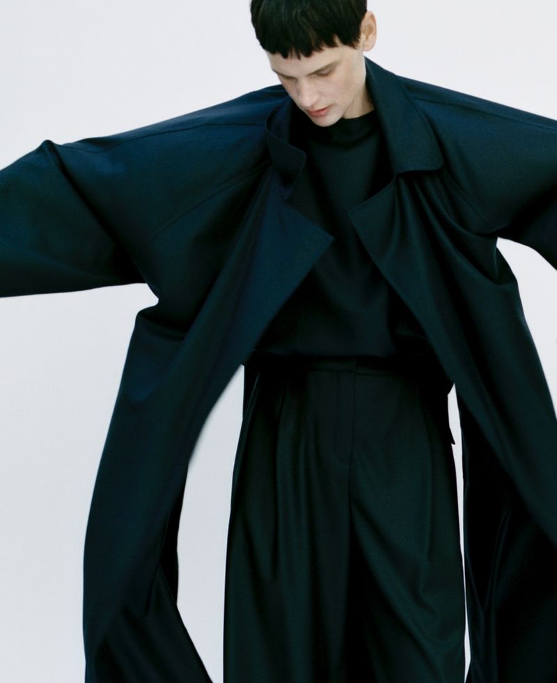 Fashion model in a dark blue, oversized coat and trousers against a white background, striking a pose with arms slightly outstretched.