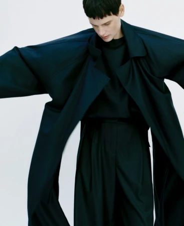 Fashion model in a dark blue, oversized coat and trousers against a white background, striking a pose with arms slightly outstretched.