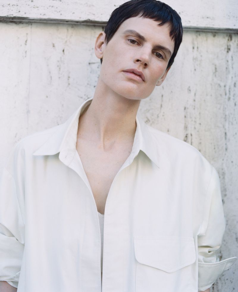 Portrait of a slender adult woman with short dark hair wearing an oversized white shirt, posing against a textured neutral wall.