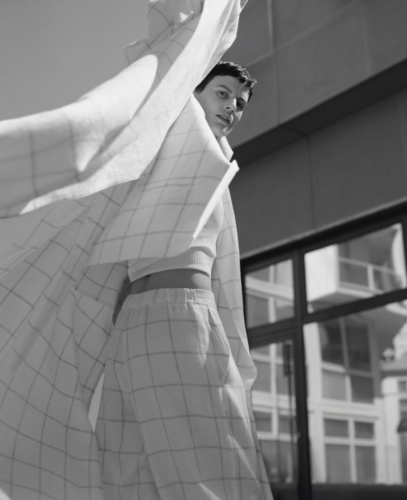 Young woman in a white plaid outfit poses outdoors near a building on a sunny day, her jacket billowing in the wind.