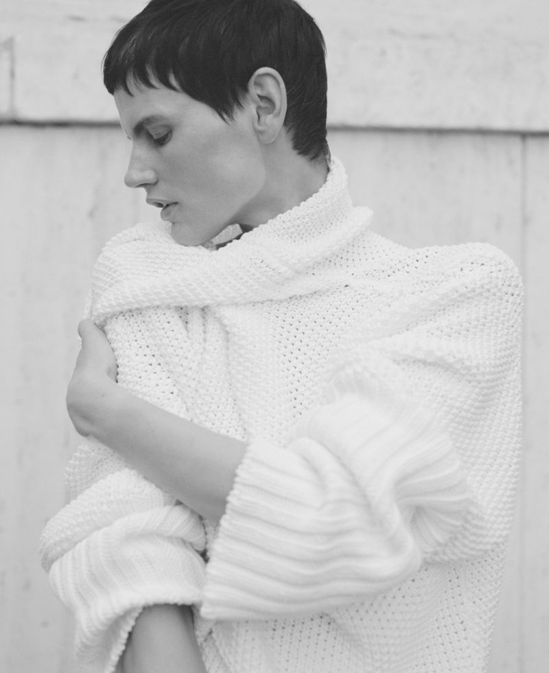 Black and white portrait of a fair-skinned woman in a white sweater, looking to the side, against a simple white background.