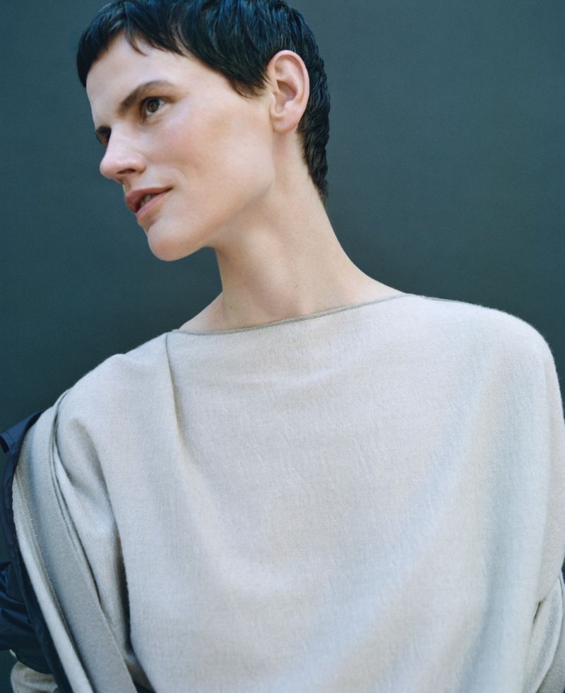 Close-up of a fair-skinned adult woman with short dark hair, wearing a light tan sweater, posing indoors against a neutral grey background.