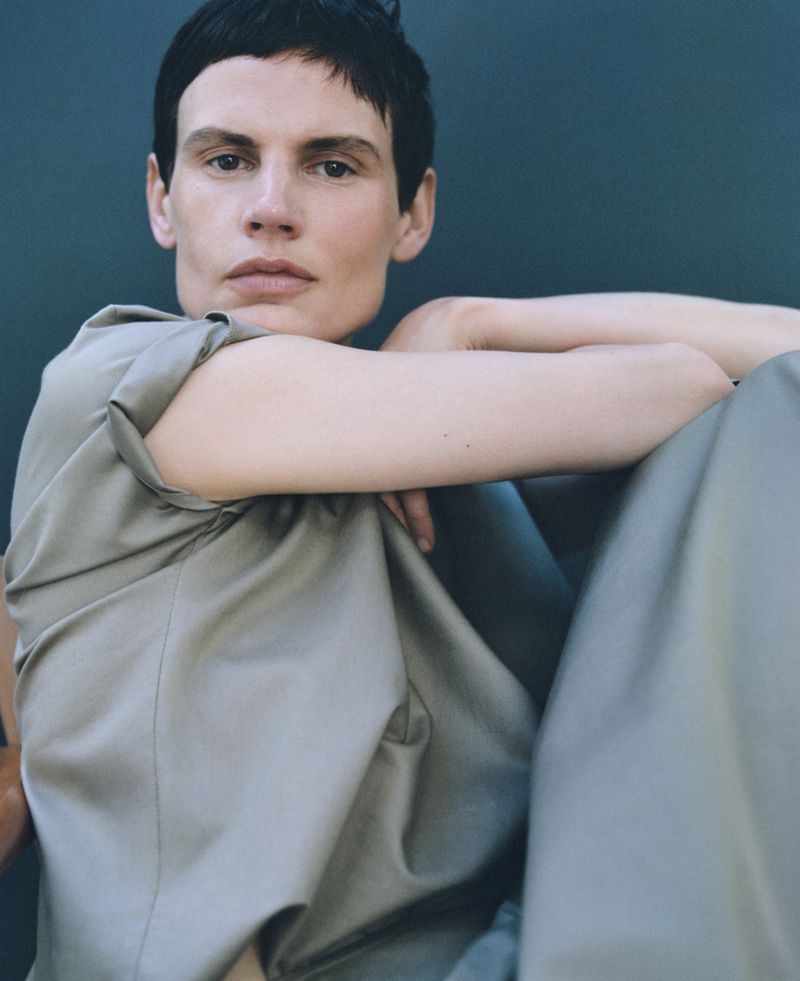Portrait of a person with short dark hair wearing a light gray outfit, set against a neutral dark gray background in a studio.