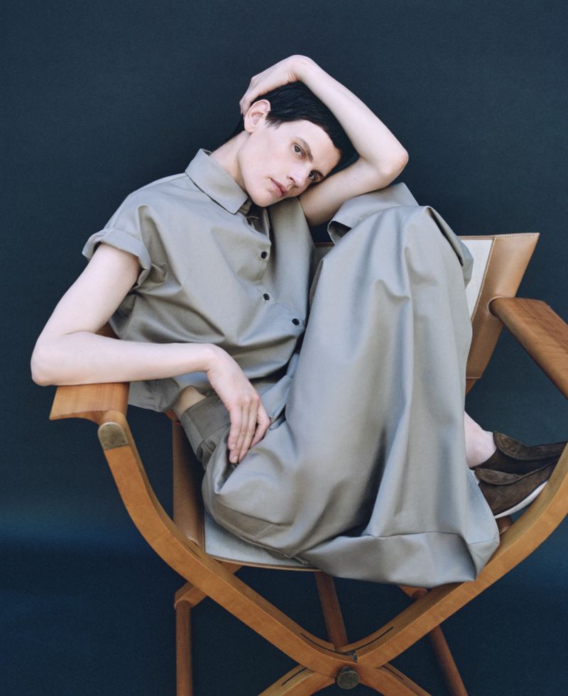 Woman in a chair wearing a grey collared shirt and pants. Neutral studio lighting, dark background. Fashion editorial style shot.