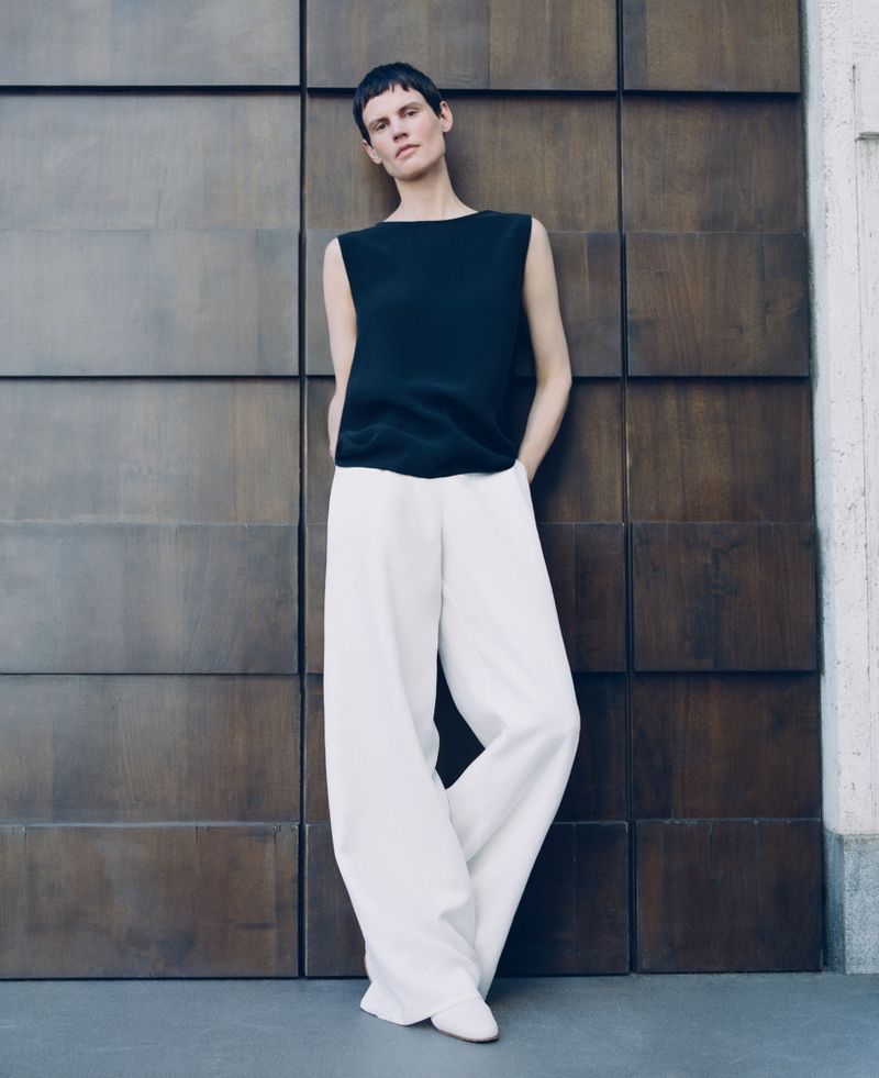 Model Saskia de Brauw in a dark sleeveless top and white wide-leg trousers leaning against dark wooden panels