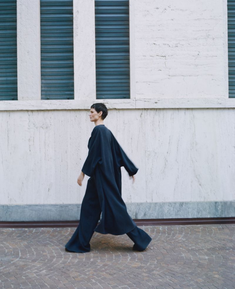 A woman in a dark blue outfit is walking outside on a patterned ground near a building with three dark horizontal rectangles.
