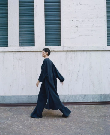 A woman in a dark blue outfit is walking outside on a patterned ground near a building with three dark horizontal rectangles.