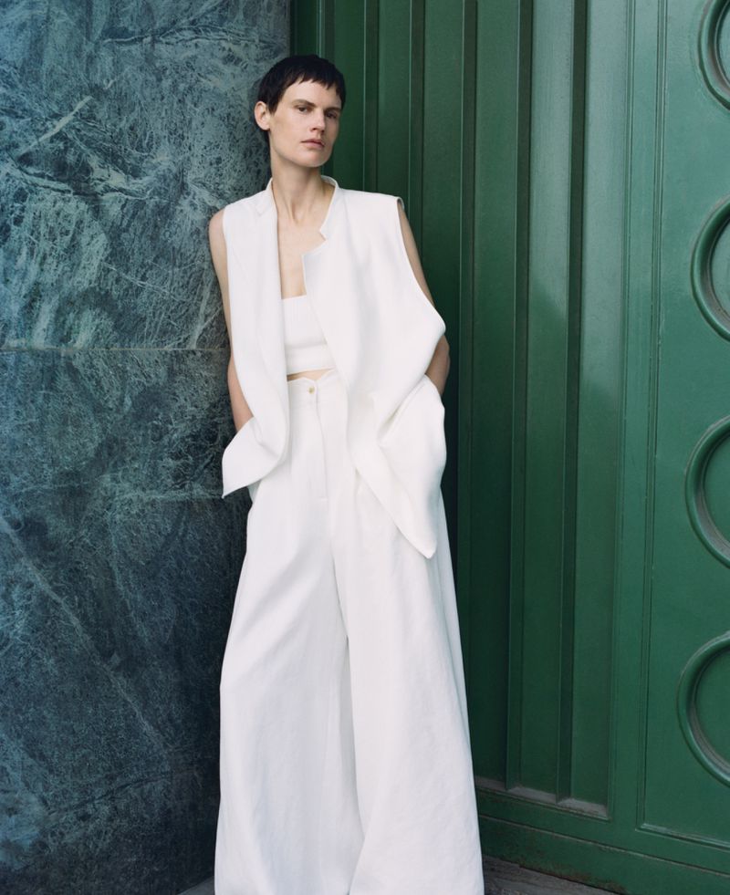A female model poses in an all-white linen vest, crop top, and wide-leg pants against a mottled blue-green marble wall and green door.