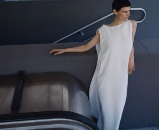 Model Saskia de Brauw in a white sleeveless dress leaning against a dark architectural wall for Dušan