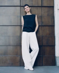 Model Saskia de Brauw in a dark sleeveless top and white wide-leg trousers leaning against dark wooden panels