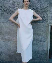 Model Saskia de Brauw in a sleeveless white midi dress posing against a textured stone wall for Dusan.