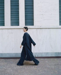Model Saskia de Brauw walking past a white building exterior wearing a dark long coat for Dusan Spring Summer 2026.