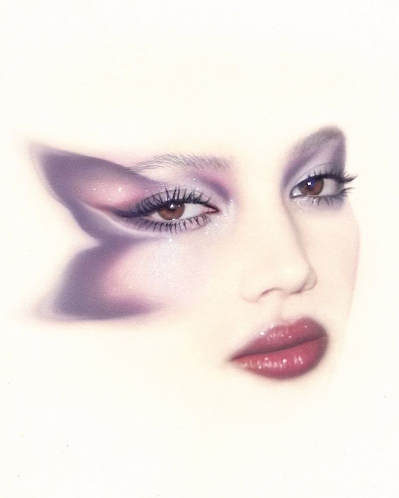 Close-up portrait of a woman with dramatic, lavender eye makeup and glossy lips, showcasing an ethereal, artistic beauty look.