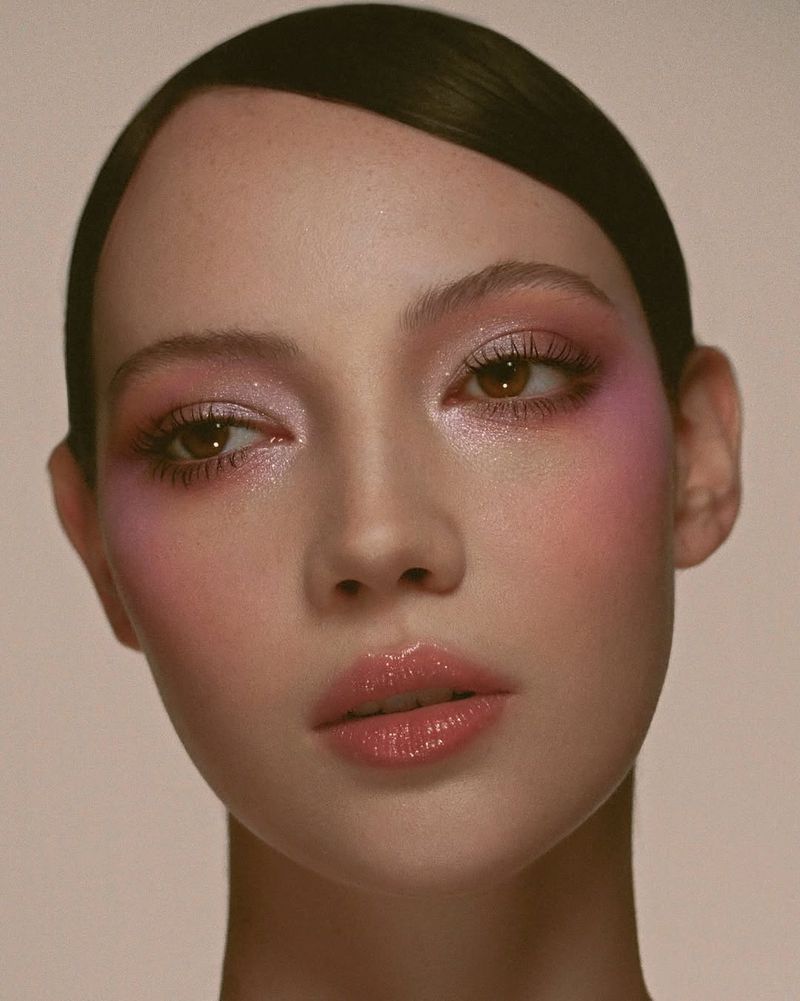 Close-up portrait of a young adult female with pink eye shadow and glossy lips, posing against a neutral-colored backdrop in a beauty shot.