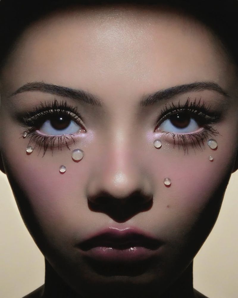 Close-up portrait of a woman with striking eyelashes and teardrop-shaped embellishments on her face, set against a neutral backdrop.