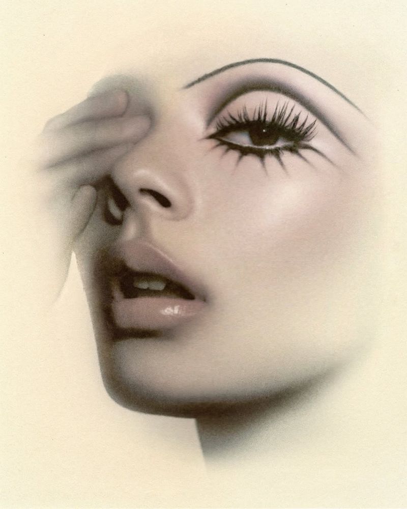 Woman with dramatic makeup and hand partially covering face, against a soft, neutral background, creating an intriguing portrait.